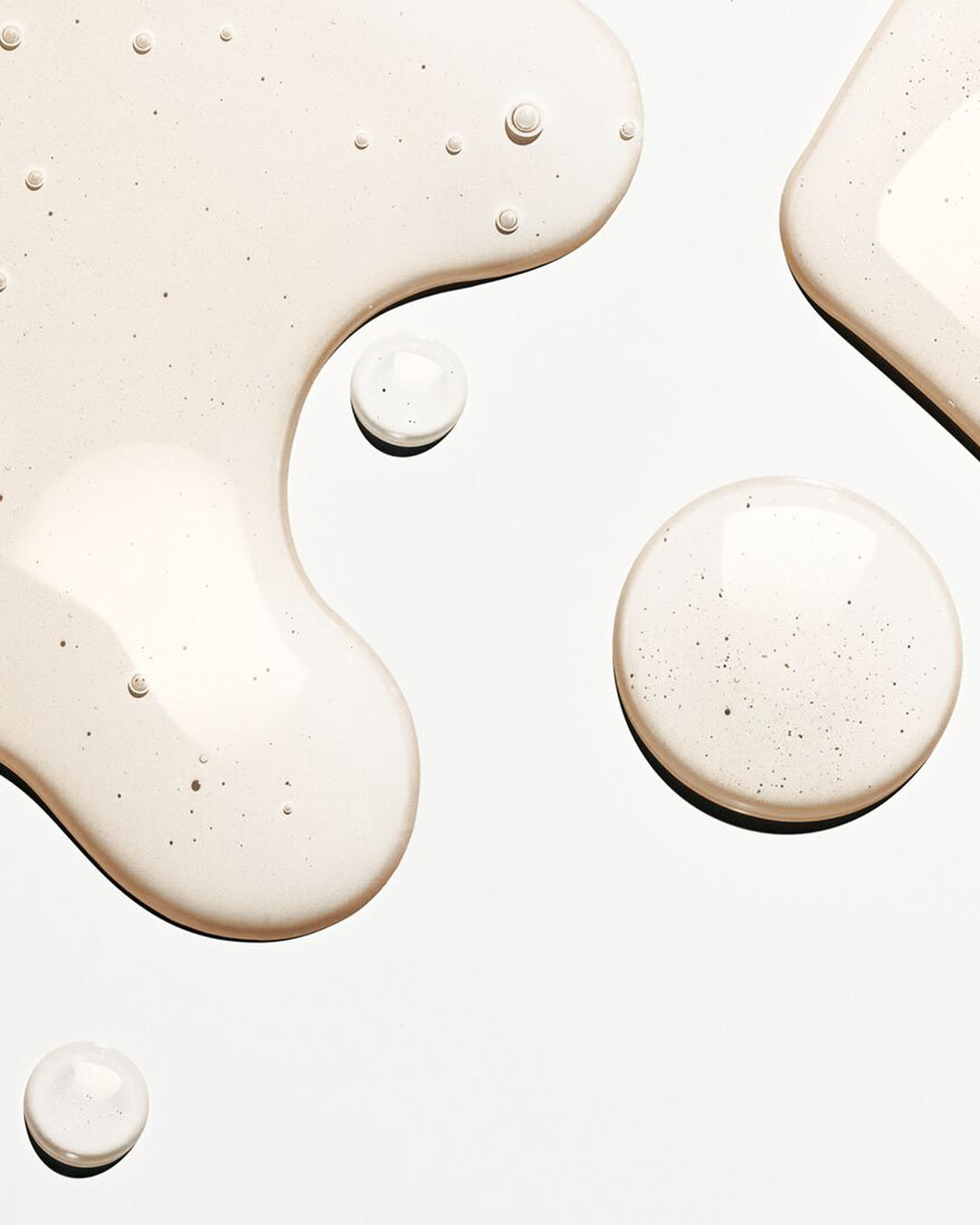 Šviesiai smėlio spalvos skysčio purslai ir lašai ant balto paviršiaus su mažais burbuliukais, sukuriančiais gaivumo ir drėkinimo pojūtį.