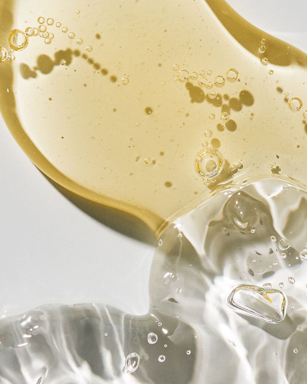 Šviesiai geltonos spalvos serumo tekstūra su burbuliukais ir skaidraus gelio tekstūra su burbuliukais ant balto paviršiaus.