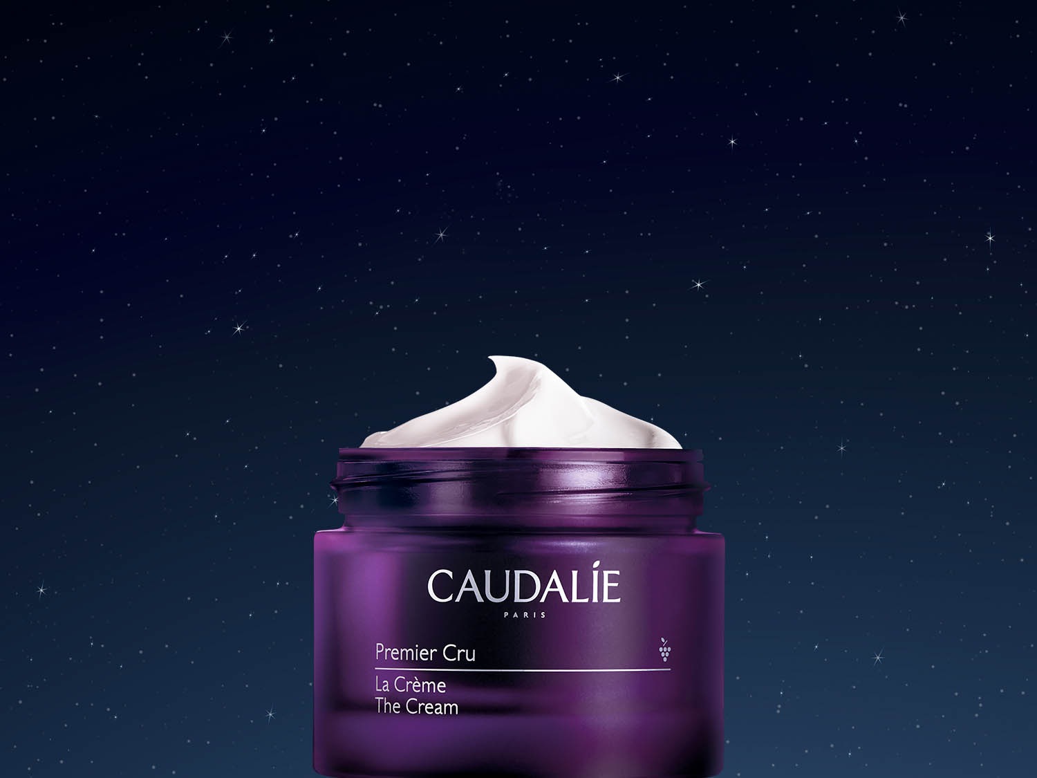 Fioletowy słoik kremu Caudalie Premier Cru La Crème na tle nocnego nieba, wypełniony białym kremem o bogatej konsystencji.