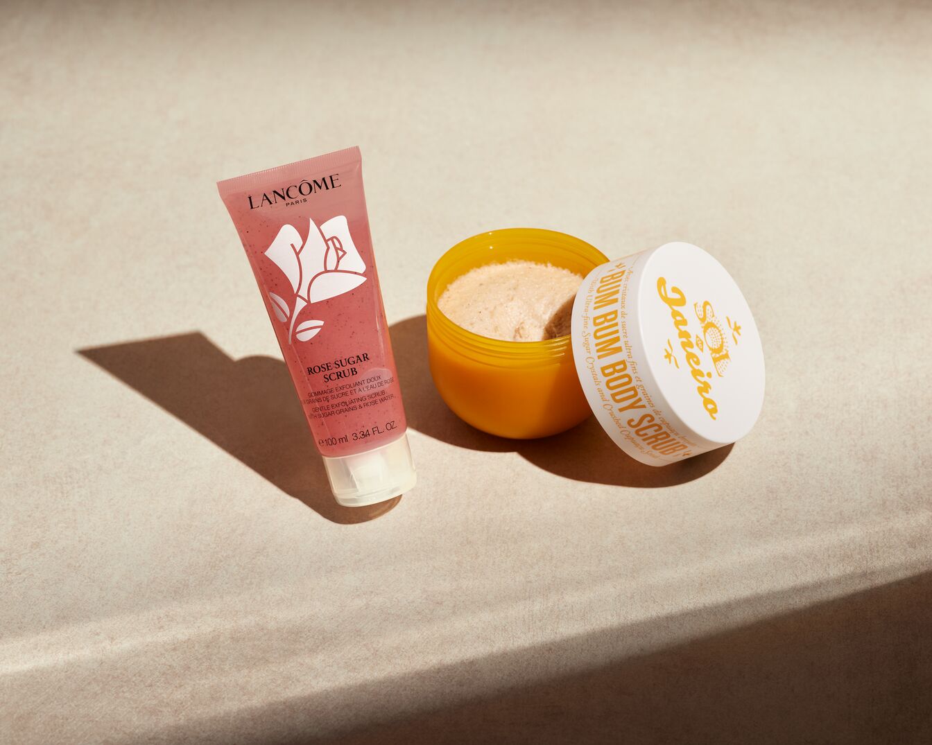 Das Lancôme Rose Sugar Scrub in einer rosa Tube mit einer weißen Rosenzeichnung daneben, daneben ein geöffneter gelber Tiegel Sol de Janeiro Bum Bum Body Scrub mit weißem Deckel.