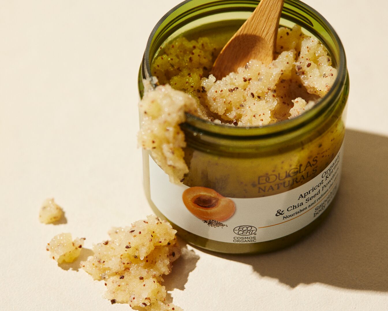 Nahaufnahme eines grünen Glastiegels von Douglas Naturals mit Aprikosen- und Chiasamen-Peeling, das mit einem Holzspatel entnommen wird, um die nährende und verfeinernde Wirkung auf die Haut zu zeigen.