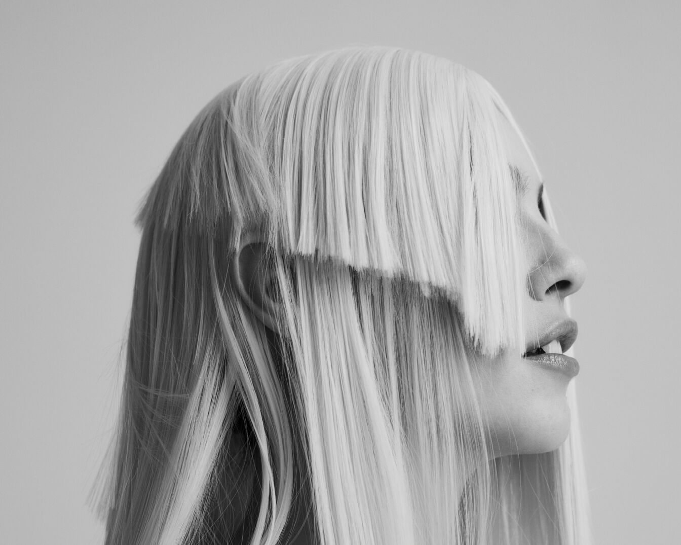 Detailní boční pohled na ženu s platinově blond parukou, jejíž ofina jí zakrývá oči a dodává jí tajemný vzhled.