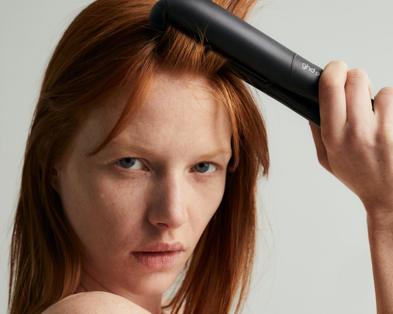 Kobieta o rudych włosach używa czarnej prostownicy GHD, aby wyprostować włosy, patrząc prosto w obiektyw.