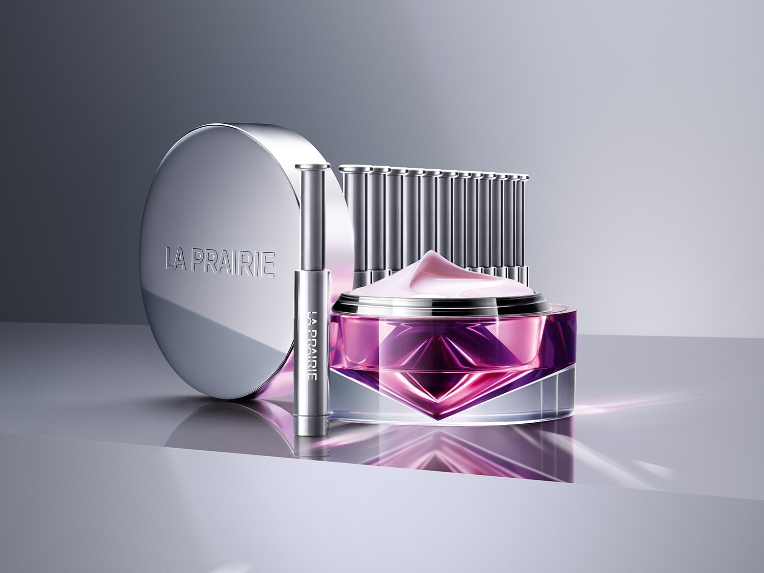 Nahaufnahme von La Prairie Hautpflegeprodukten, darunter eine violette Creme in einem transparenten Tiegel, ein silberner Lippenstift und eine Dose mit silbernem Deckel, die auf einer glänzenden Oberfläche stehen.