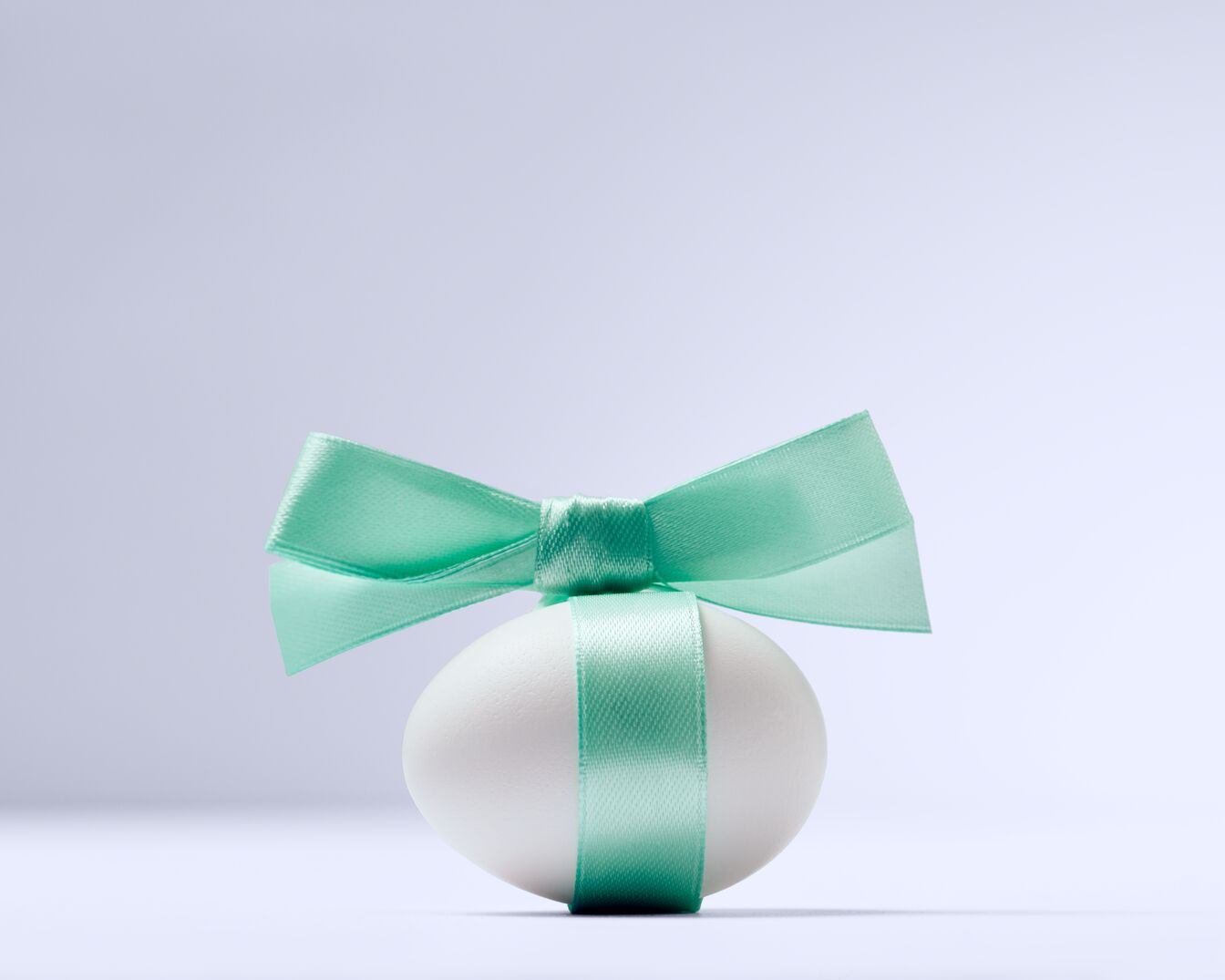 Białe jajko wielkanocne owinięte turkusową wstążką i kokardą na jasnym tle.