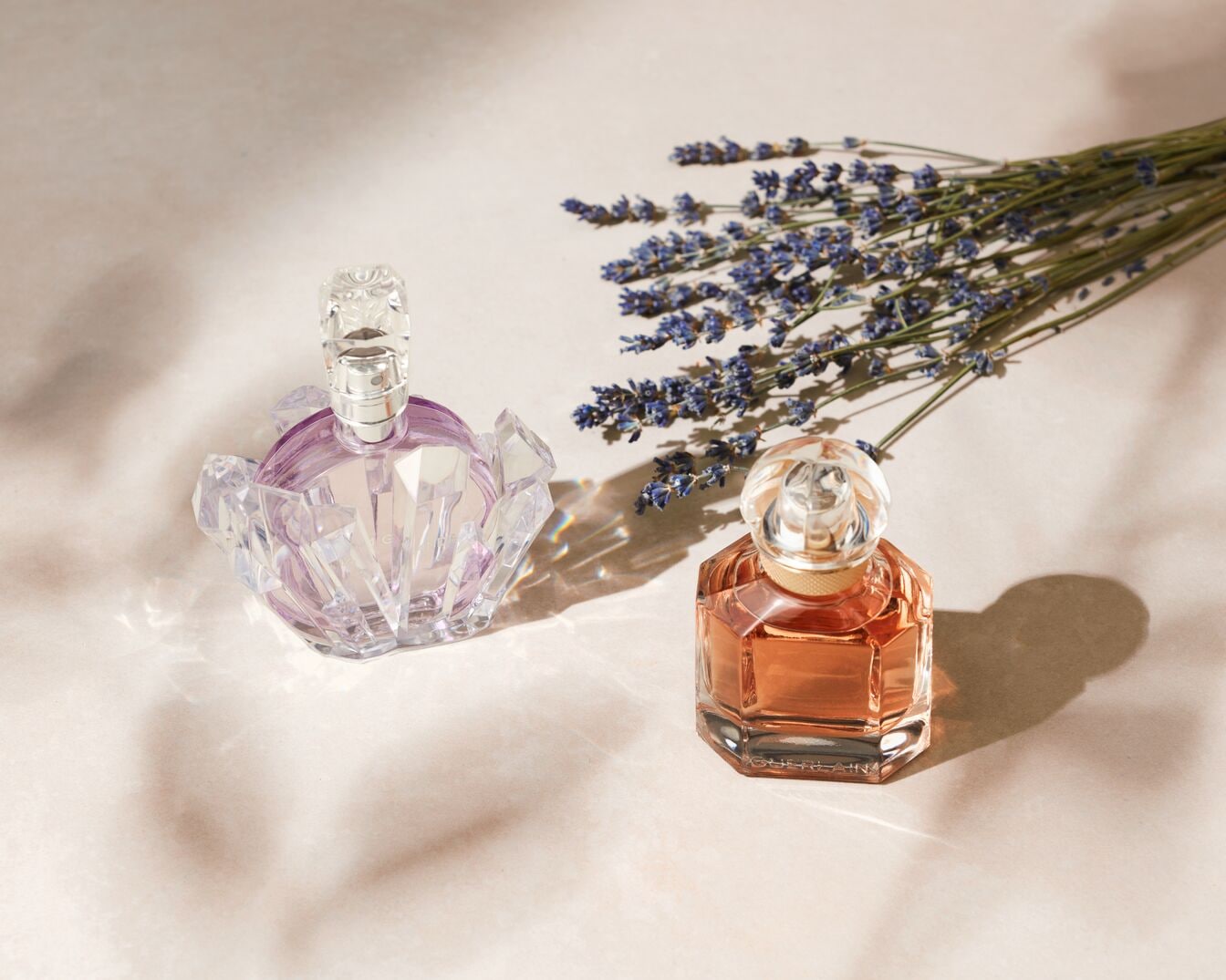 Zwei Parfümflaschen, eine in einem klaren, facettierten Flakon mit einem Hauch von Lila und die andere in einem bernsteinfarbenen, achteckigen Flakon, liegen neben einem Strauß Lavendel.