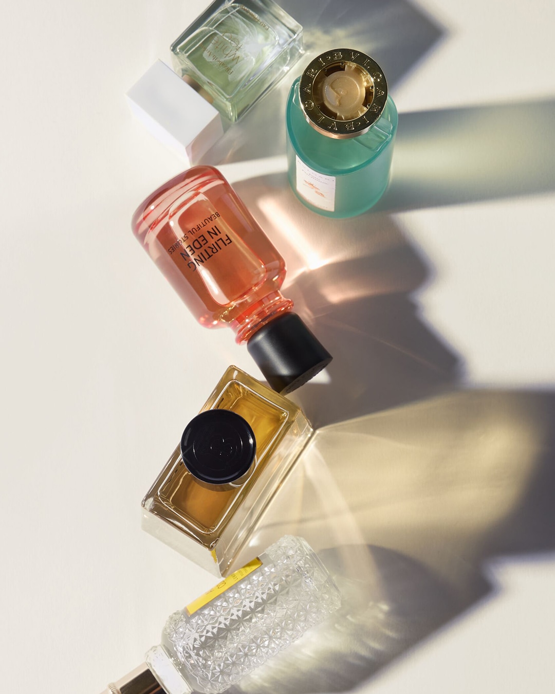 Verschiedene Parfümflakons in Grün-, Pfirsich- und Goldtönen liegen auf einer hellen Oberfläche und werfen lange Schatten.