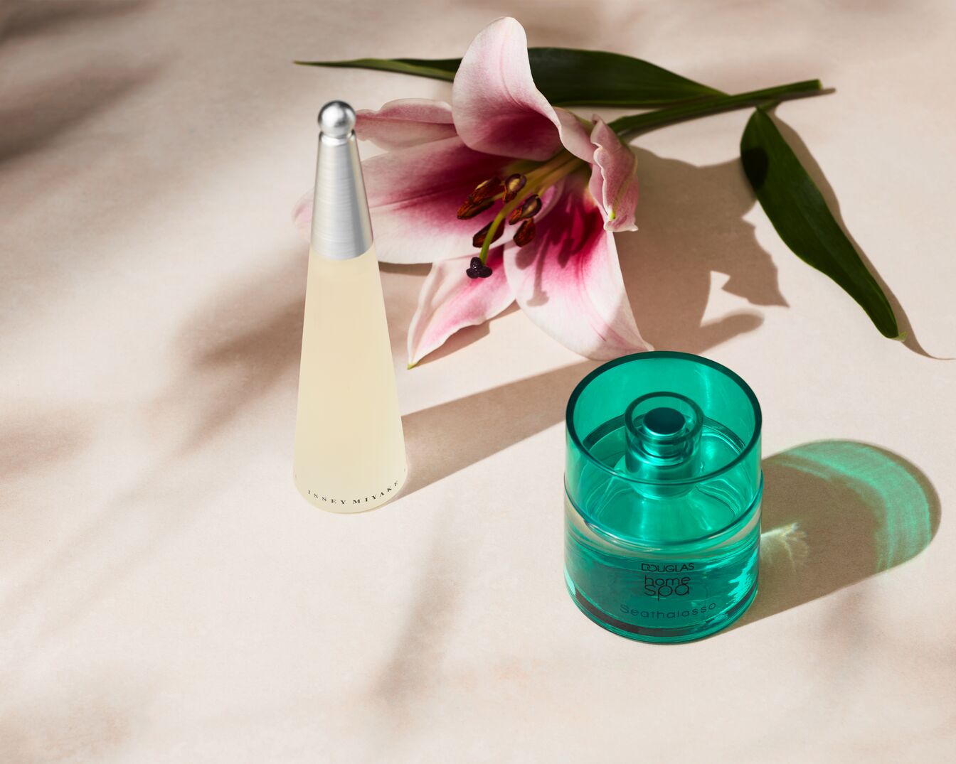 En la imagen se aprecia un frasco de perfume Issey Miyake con forma cónica y tapa plateada, junto a un difusor verde azulado de Douglas Home Spa, ambos acompañados de una delicada flor de lirio rosada.