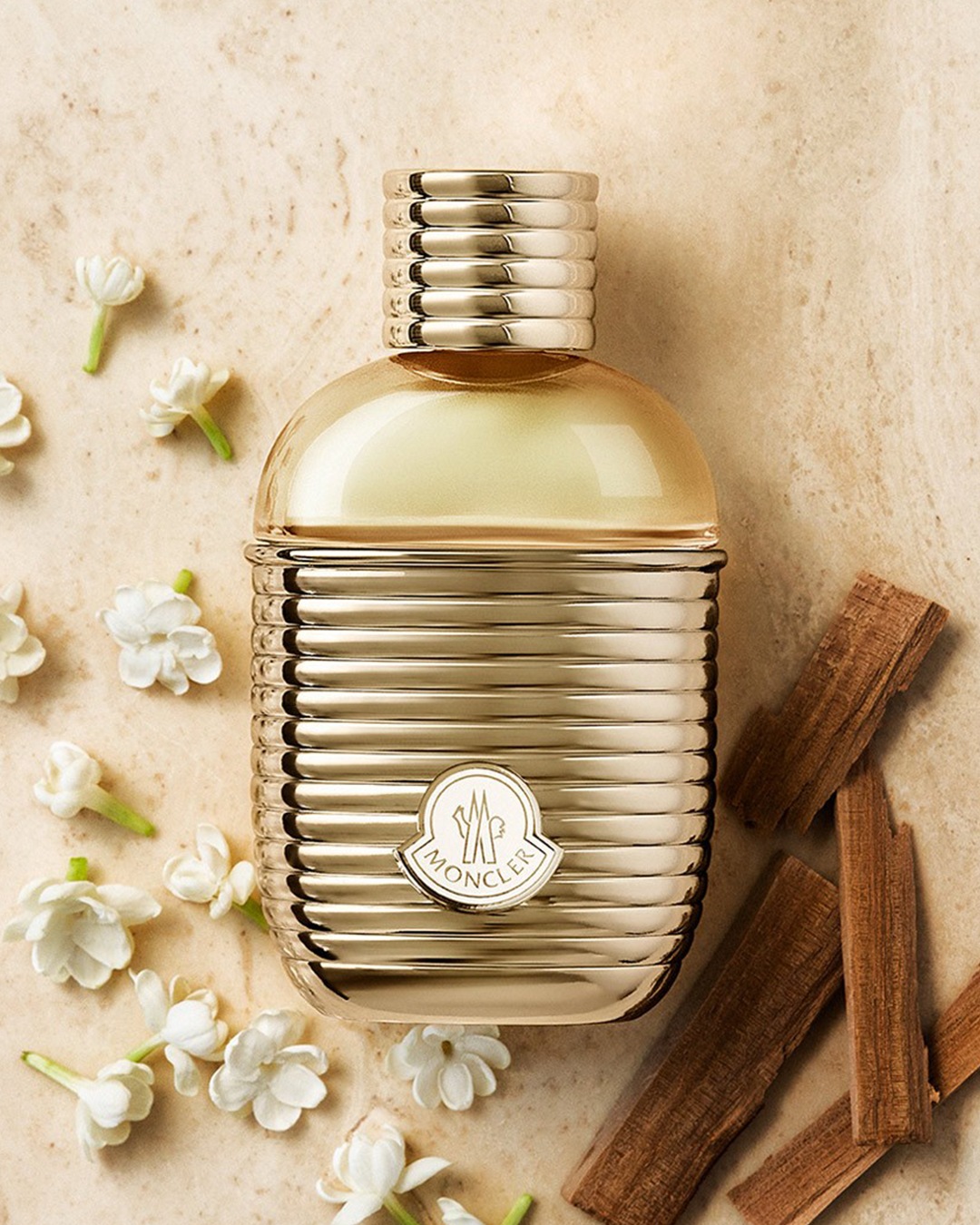 Gouden Moncler parfumfles met ribbels, omringd door witte bloemen en houten stokjes, voor een luxe en verfijnde geur.