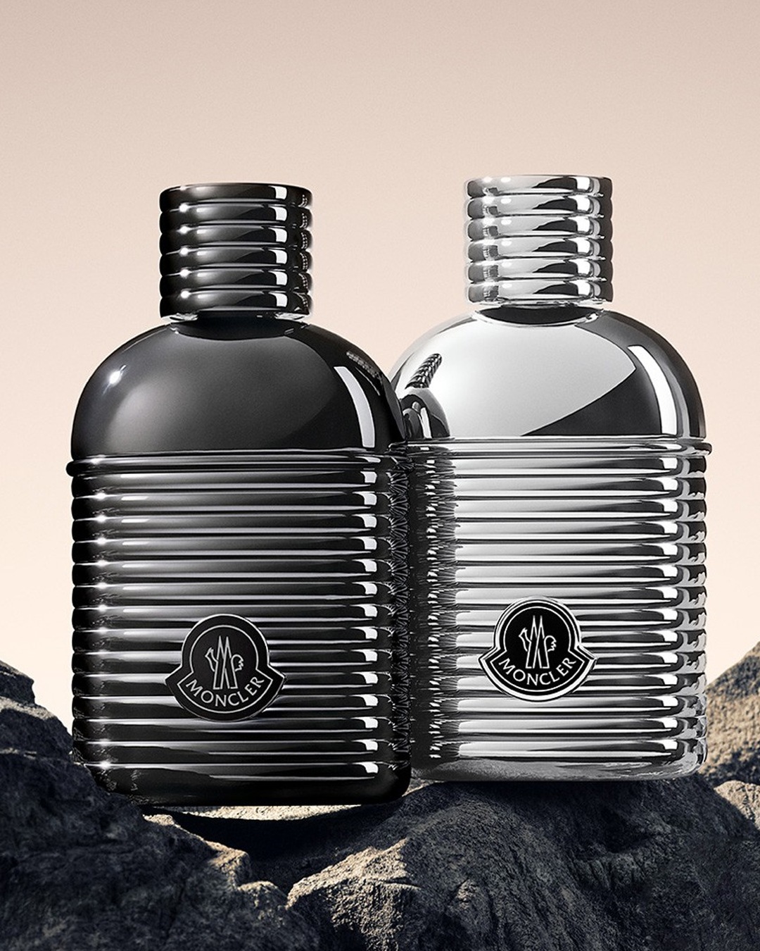Twee Moncler parfumflessen, een in zwart en een in zilver, staan op een rotsachtige ondergrond, met het Moncler logo prominent weergegeven op beide flessen.