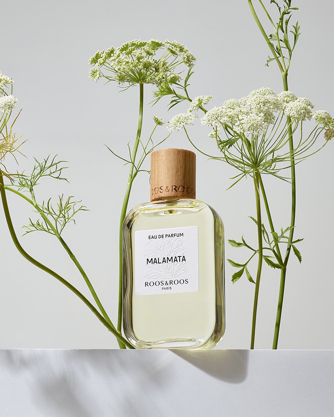 Découvrez le parfum MALAMATA de ROOS&ROOS Paris, une fragrance élégante présentée dans un flacon transparent avec un bouchon en bois, entouré de délicates fleurs blanches.
