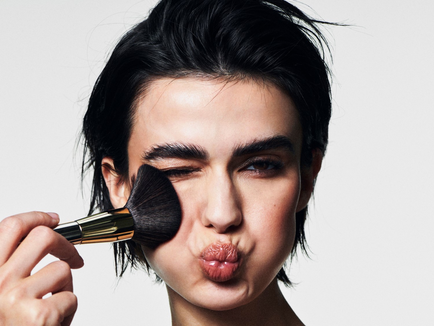 Eine Frau mit dunklen Haaren und einem goldenen Make-up-Pinsel, die sich schminkt und dabei kokett in die Kamera blickt.