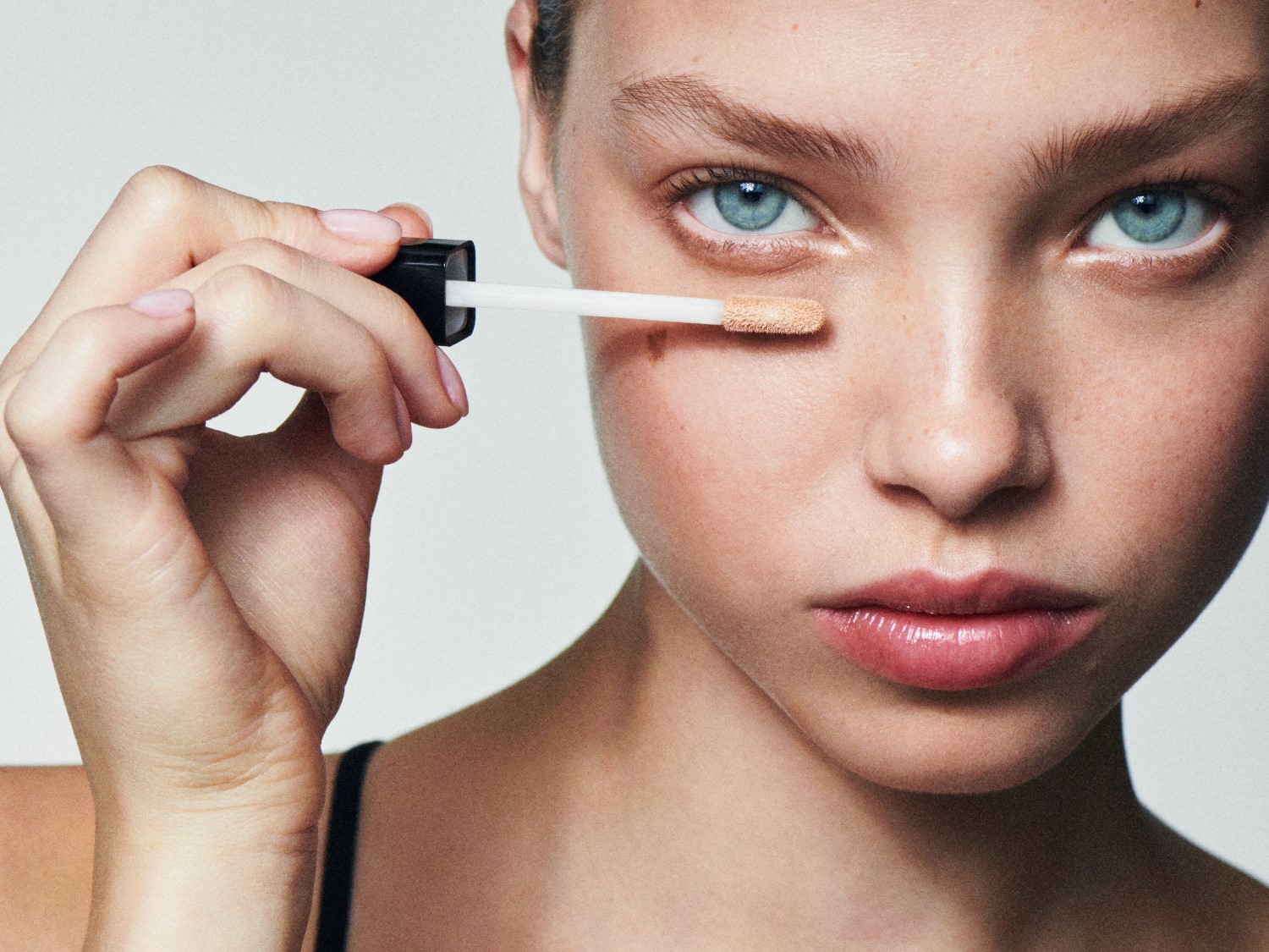 Nahaufnahme eines jungen Models mit strahlend blauen Augen, das mit einem Applikator Concealer unter dem Auge aufträgt, um einen ebenmäßigen Teint zu erzielen.