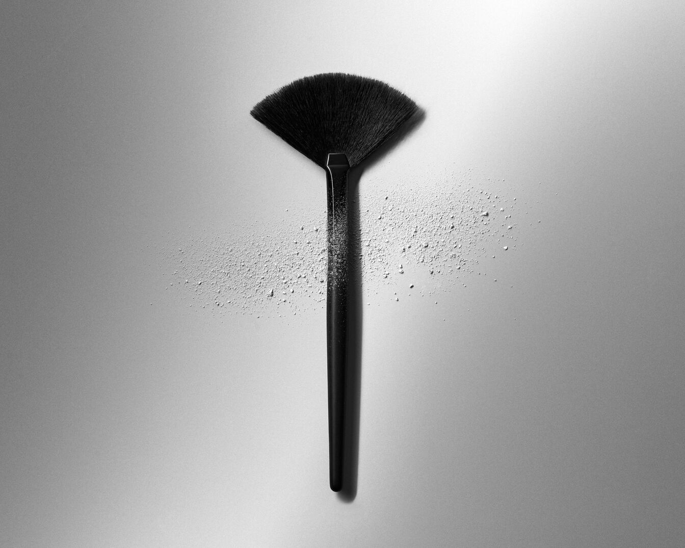 Ein schwarzer Fächerpinsel liegt auf einem grauen Untergrund, daneben ist loser, heller Puder zu sehen.
