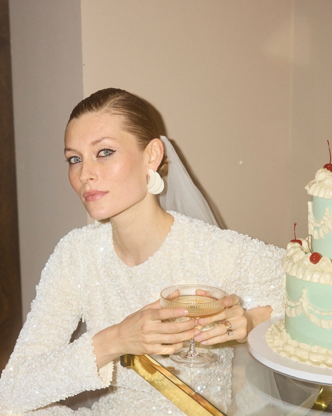 Nuotaka baltu blizgančiu kostiumėliu laiko taurę šampano prie žalios vestuvinio torto su vyšniomis.