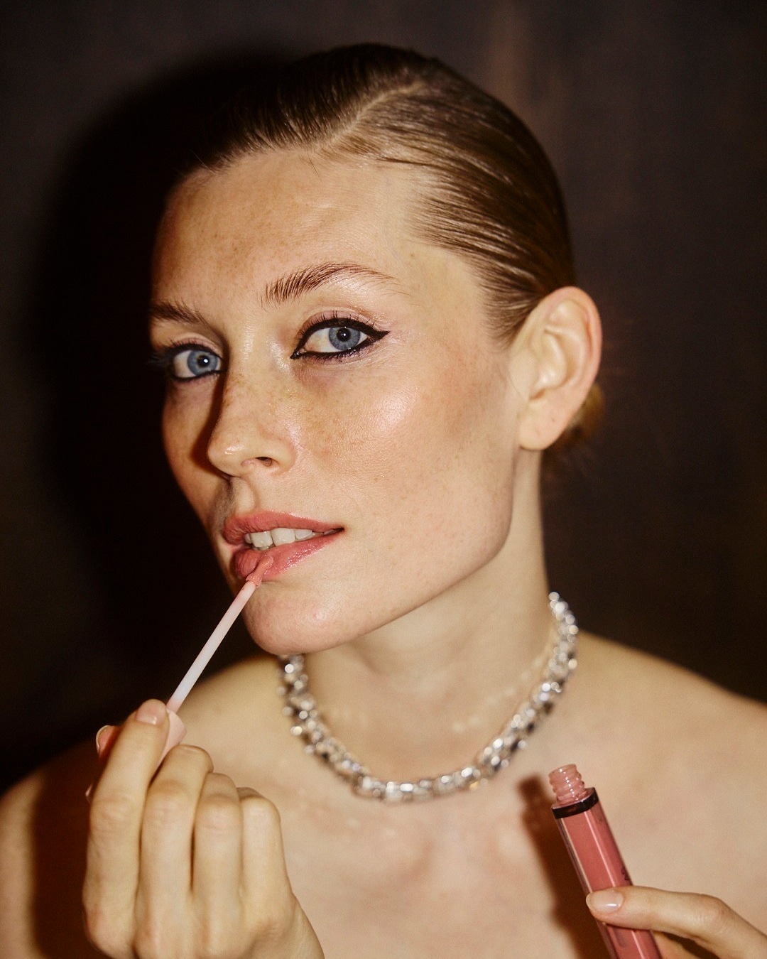 Šviesiaplaukė moteris tepa lūpų blizgesį, pabrėždama savo natūralų grožį ir eleganciją su sidabriniu vėriniu.
