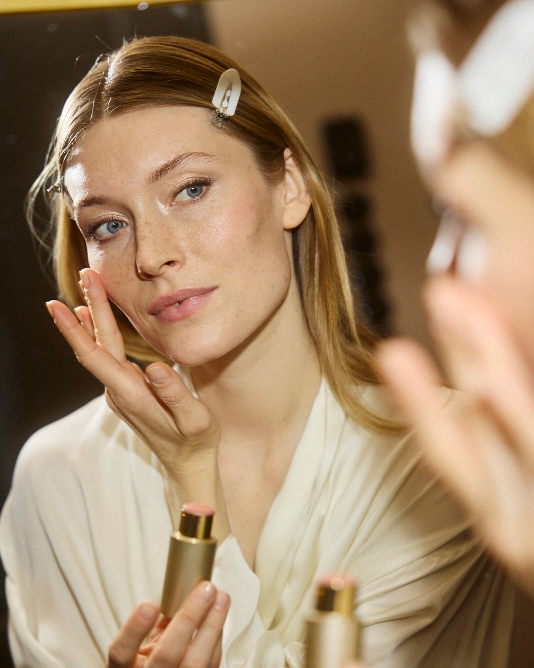 Šviesiaplaukė moteris tepa skaistalus ant skruosto, laikydama rankoje auksinės spalvos tūbelę su rausvu dangteliu, kad oda būtų skaisti.