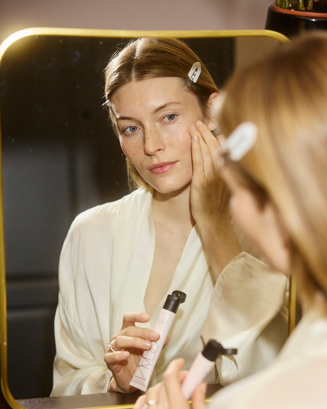 Moteris, vilkinti chalatą, tepa veidą NARS produktu, atsispindėdama veidrodyje.