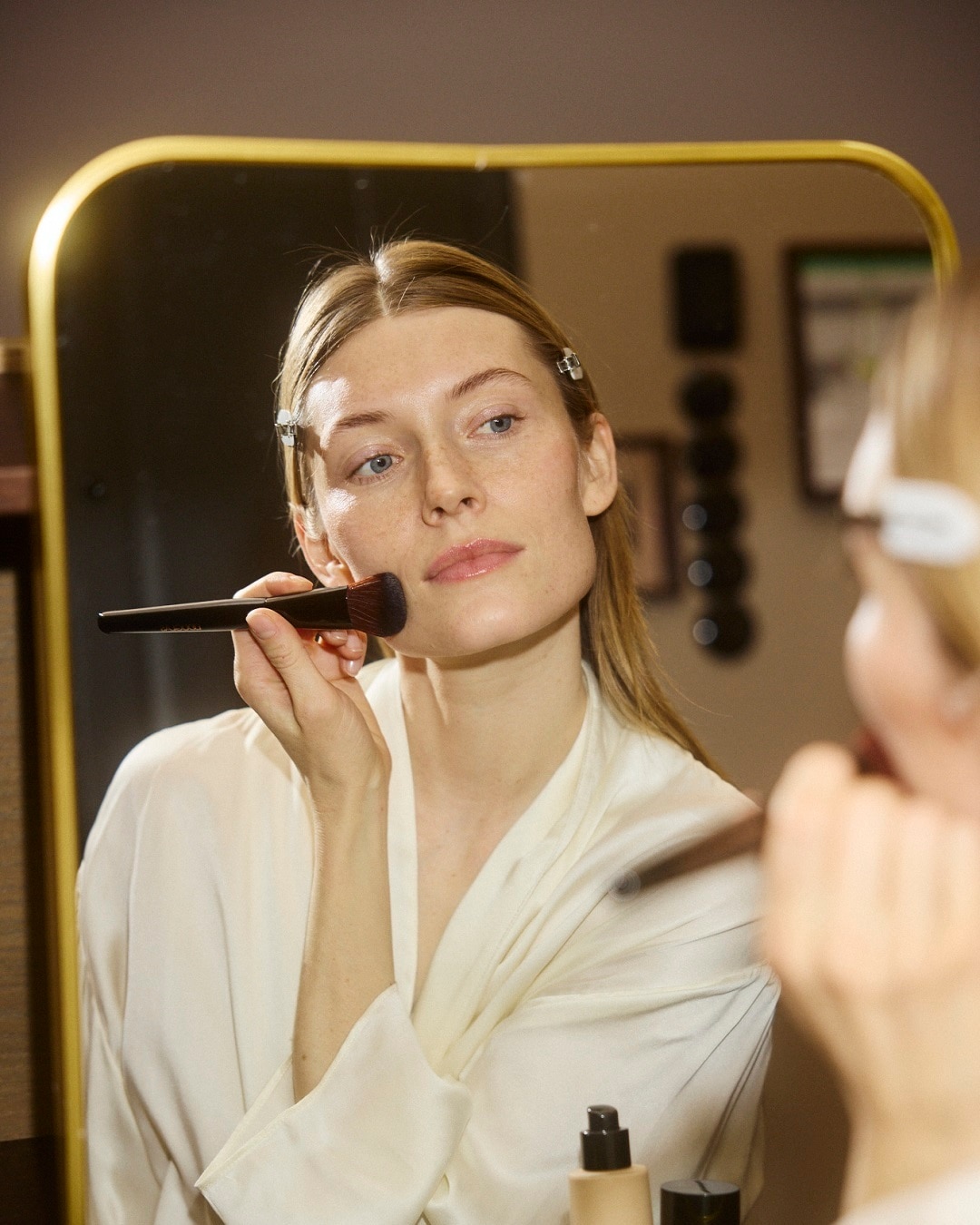 Moteris, vilkinti chalatą, veidrodyje tepa makiažą teptuku, kad sukurtų nepriekaištingą išvaizdą.
