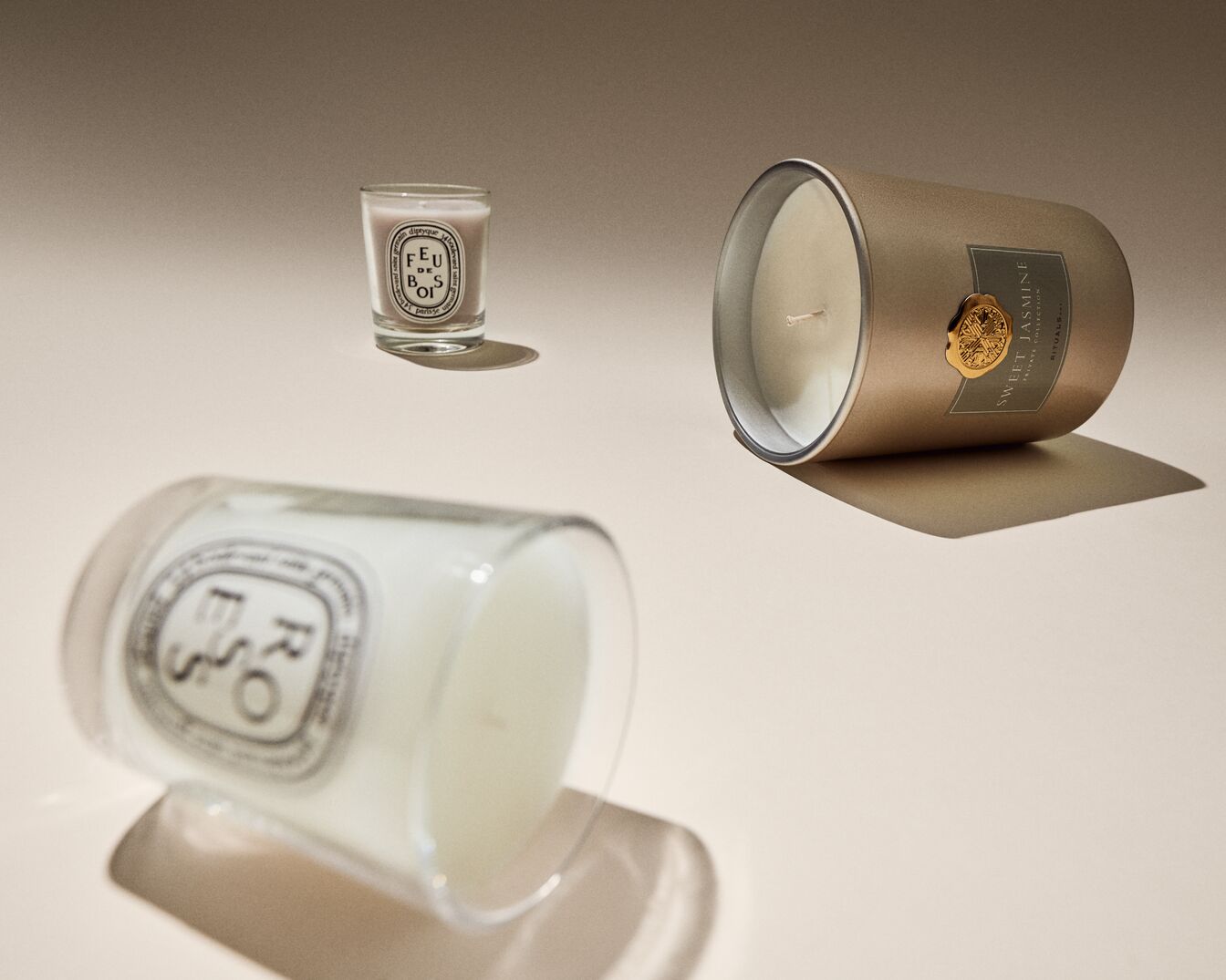 Eine Nahaufnahme von drei Duftkerzen unterschiedlicher Größe und Form, darunter eine "Feu de Bois"-Kerze in einem Glasbehälter und eine "Sweet Jasmine"-Kerze in einem goldenen Behälter.