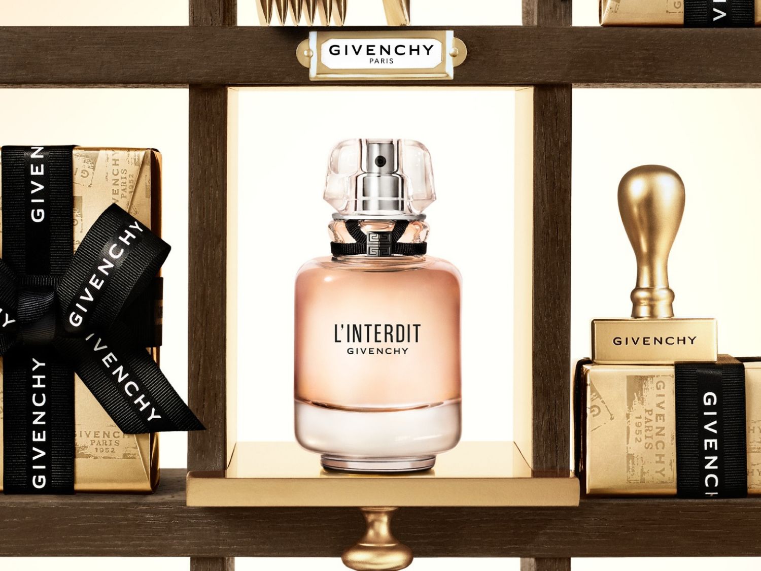 Ein Givenchy L'Interdit Parfümflakon mit rosafarbener Flüssigkeit und einem transparenten Verschluss steht auf einem goldenen Regal, umgeben von goldenen Geschenkboxen mit schwarzen Bändern, die das Givenchy-Logo tragen.