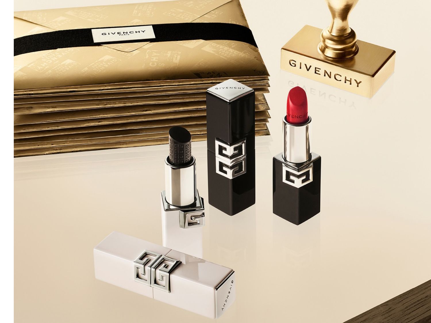 Drei Givenchy Lippenstifte in Schwarz und Weiß, einer davon in leuchtendem Rot, sind zusammen mit einem goldenen Stempel und einem Stapel goldener Umschläge auf einer cremefarbenen Oberfläche arrangiert.