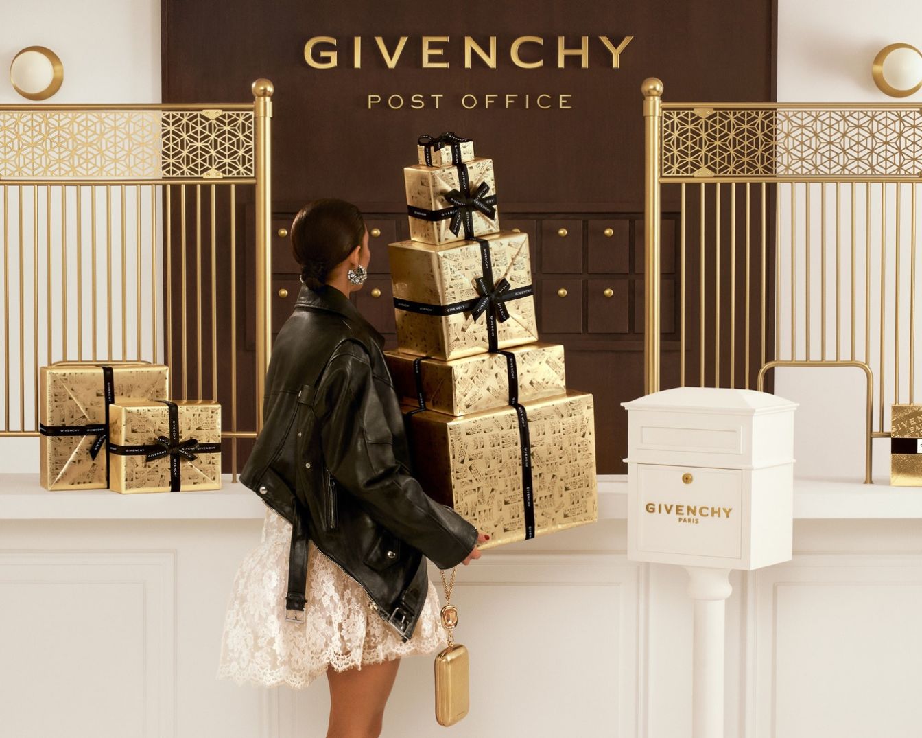 Eine Frau in einer schwarzen Lederjacke und einem weißen Spitzenkleid steht vor einem Givenchy Post Office-Schild und hält einen Stapel goldener Geschenke mit schwarzen Bändern.
