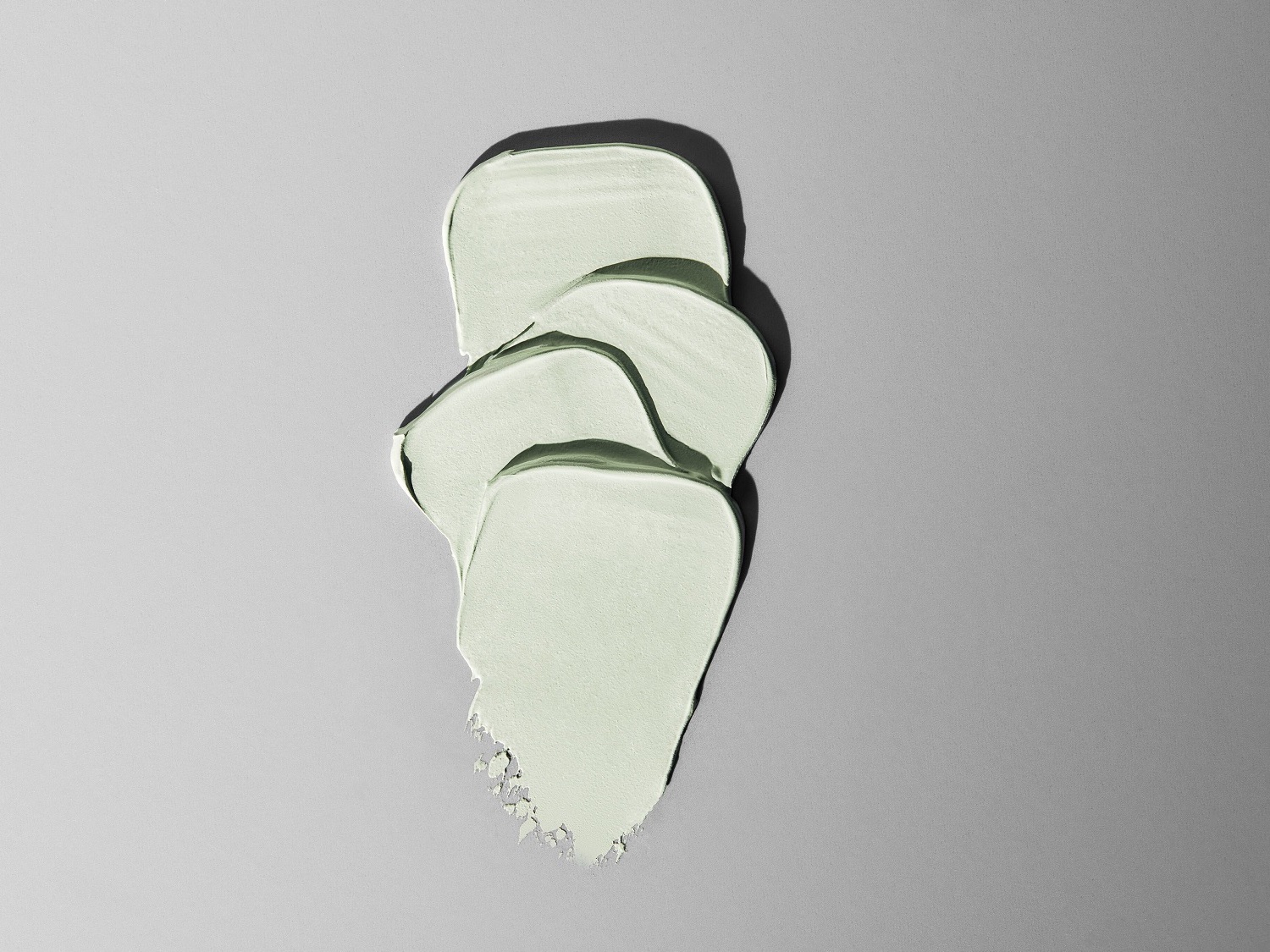 Nahaufnahme eines hellgrünen Cremeprodukts, das in dicken Schichten auf einer grauen Oberfläche aufgetragen wurde und eine glatte, fast pastellartige Textur zeigt.