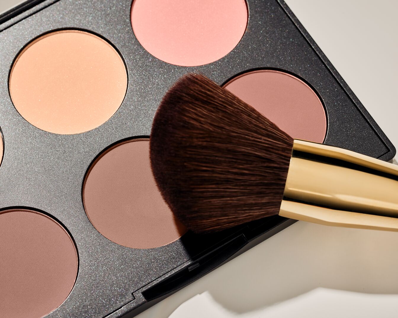 Nahaufnahme einer Make-up-Palette mit verschiedenen Rouge- und Bronzer-Farben, ergänzt durch einen Pinsel mit goldenem Griff.