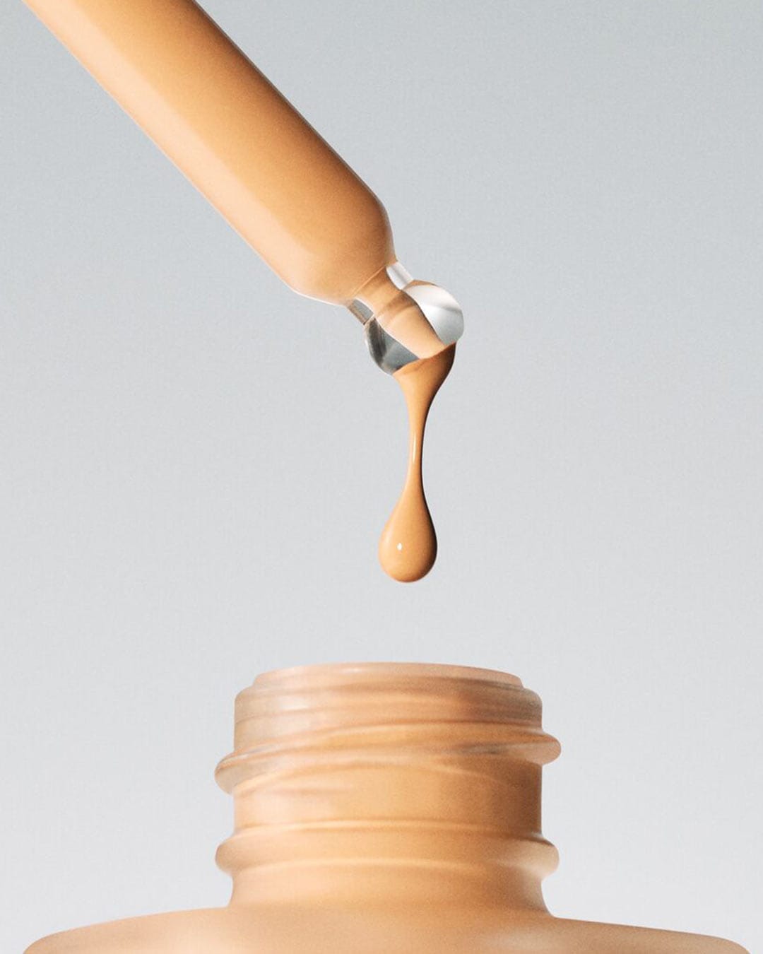 Ein Tropfen beiger Foundation fällt aus einer Pipette in eine geöffnete Flasche, um die flüssige Textur und den Farbton des Produkts hervorzuheben.