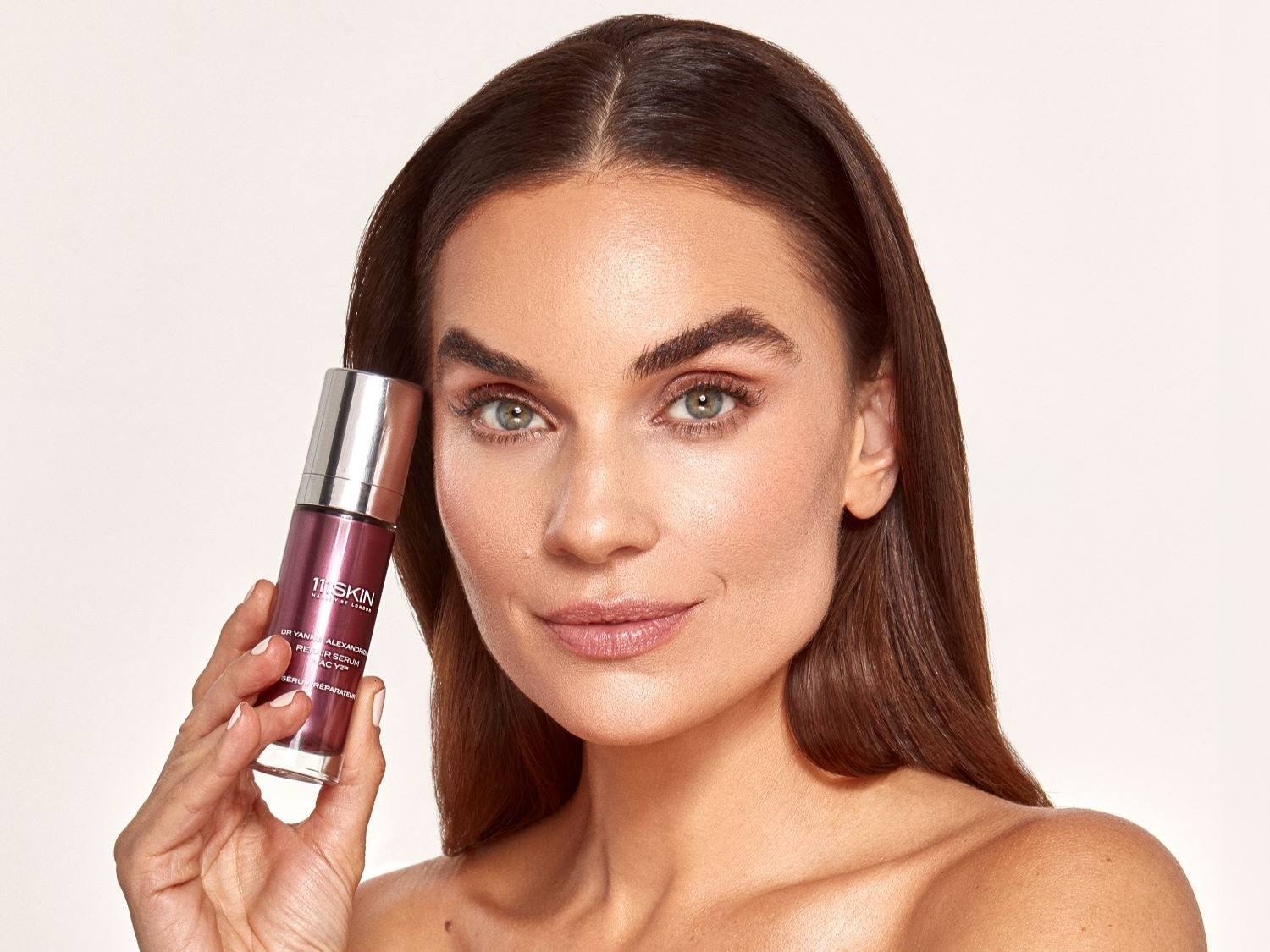 Eine Frau mit braunen Haaren hält eine Serumflasche von 111SKIN in der Hand, die Haut wirkt strahlend und gepflegt.