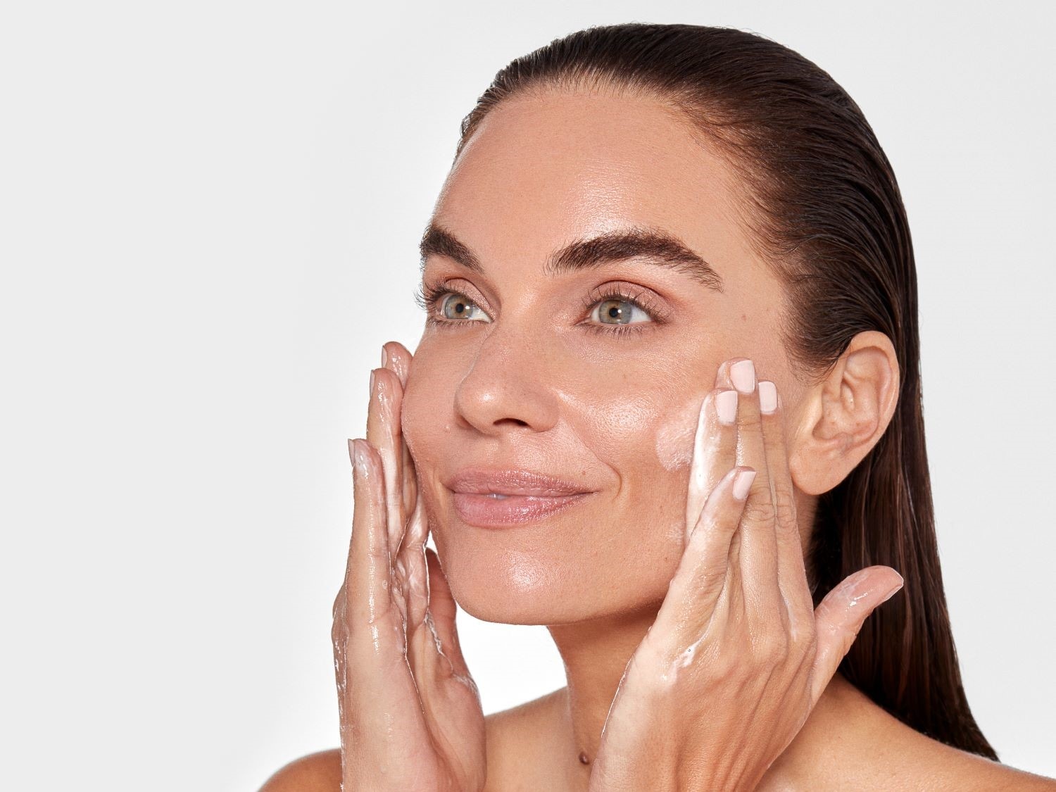 Eine Frau mit hellbraunem, zurückgekämmtem Haar massiert eine weiße Creme mit beiden Händen in ihr Gesicht ein.