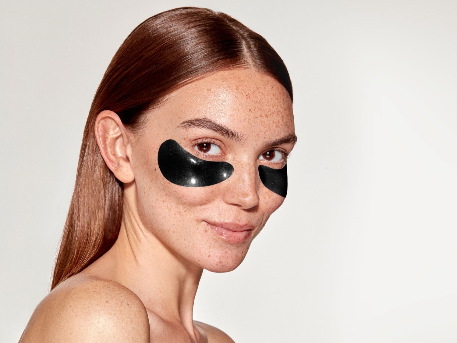 Nahaufnahme einer Frau mit Sommersprossen und glattem, braunem Haar, die schwarze Augenpads unter den Augen trägt und lächelt.