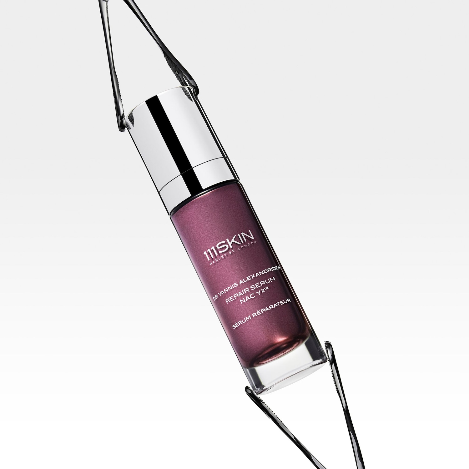 Nahaufnahme einer Serumflasche von 111Skin mit silbernem Verschluss und durchsichtigem Behälter, der das rosafarbene Serum zeigt, gehalten von zwei schwarzen Pinzetten.