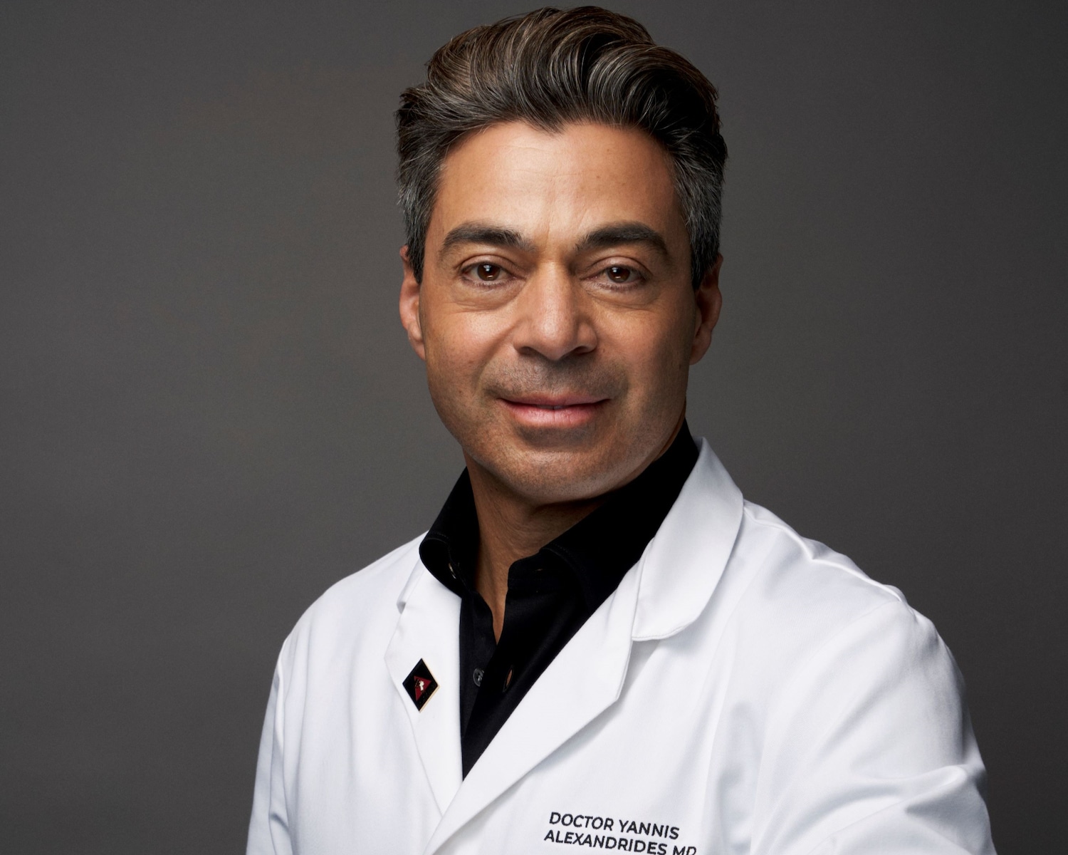 Porträt von Doktor Yannis Alexandrides, der einen weißen Arztkittel über einem schwarzen Hemd trägt und ein freundliches Lächeln zeigt.