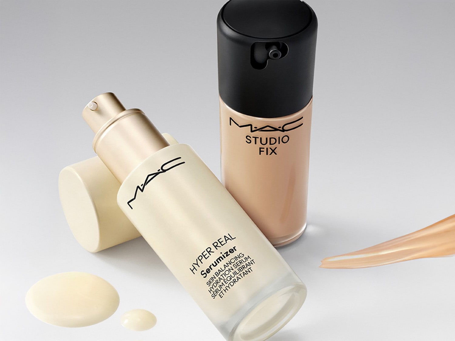 Dwa produkty MAC do makijażu: Hyper Real Serumizer w białej butelce z pompką i Studio Fix w beżowej butelce z czarną nakrętką, obok kropla serum i smugą podkładu.