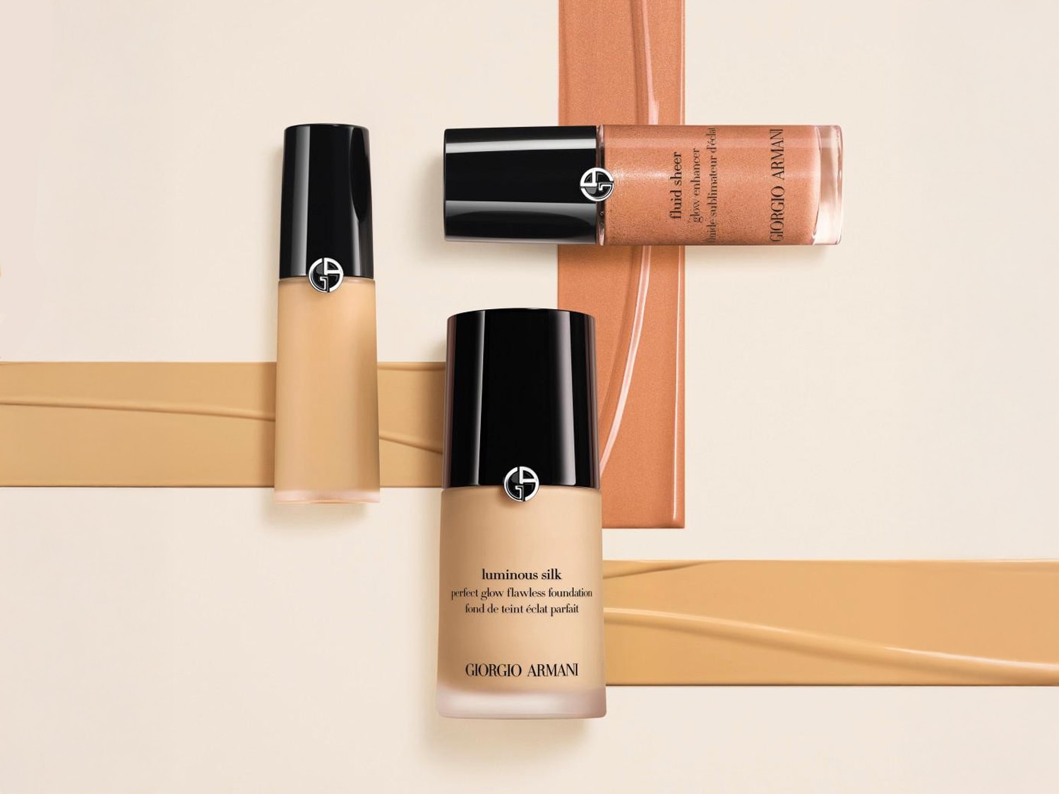 Trzy butelki podkładu Giorgio Armani Luminous Silk i Fluid Sheer Glow Enhancer z czarnymi zakrętkami na beżowym tle z paskami podkładu w różnych odcieniach.