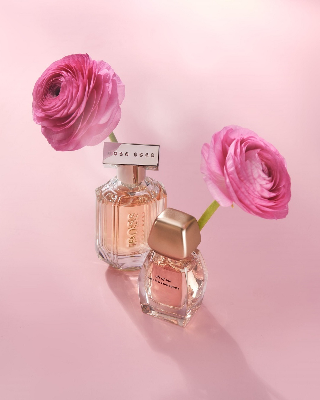 Dva flakóny parfumov Hugo Boss a Narciso Rodriguez sú umiestnené medzi ružovými kvetmi na ružovom pozadí, čo vytvára jemnú a elegantnú kompozíciu.