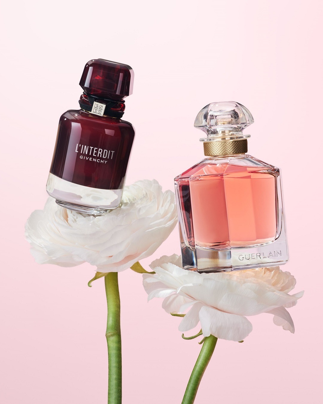 Dva flakóny parfumov Givenchy L'Interdit a Guerlain stoja na bielych kvetoch s ružovým pozadím.