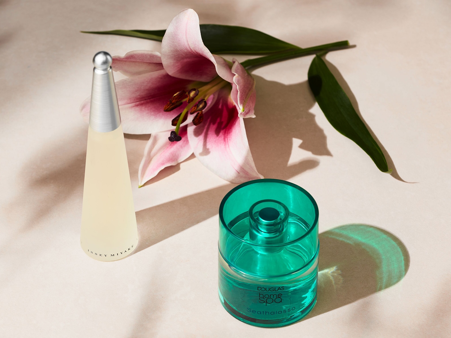 Présentation de deux parfums : un flacon conique jaune clair avec un bouchon argenté et un diffuseur d'ambiance turquoise, agrémentés d'un lys rose.