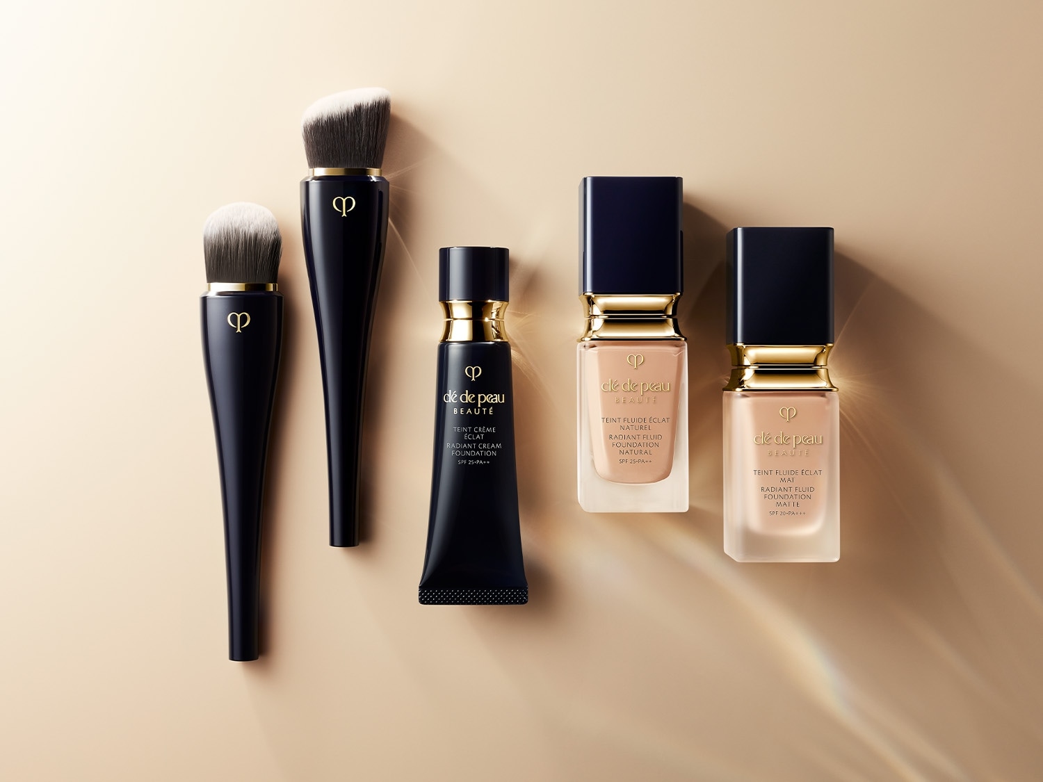 Dos brochas de maquillaje negras, un tubo de base en crema y dos frascos de base líquida de Clé de Peau Beauté sobre un fondo beige.