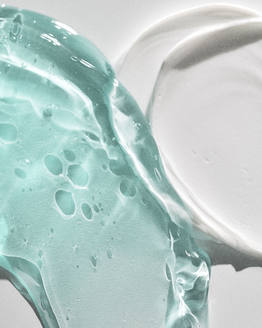 Detailní záběr na průsvitný modrozelený gel a bílý krém, které ukazují texturu a konzistenci kosmetických produktů.