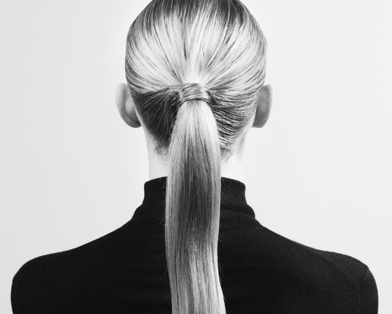 In bianco e nero, una donna con i capelli biondi raccolti in una coda di cavallo bassa indossa un dolcevita nero, vista da dietro.