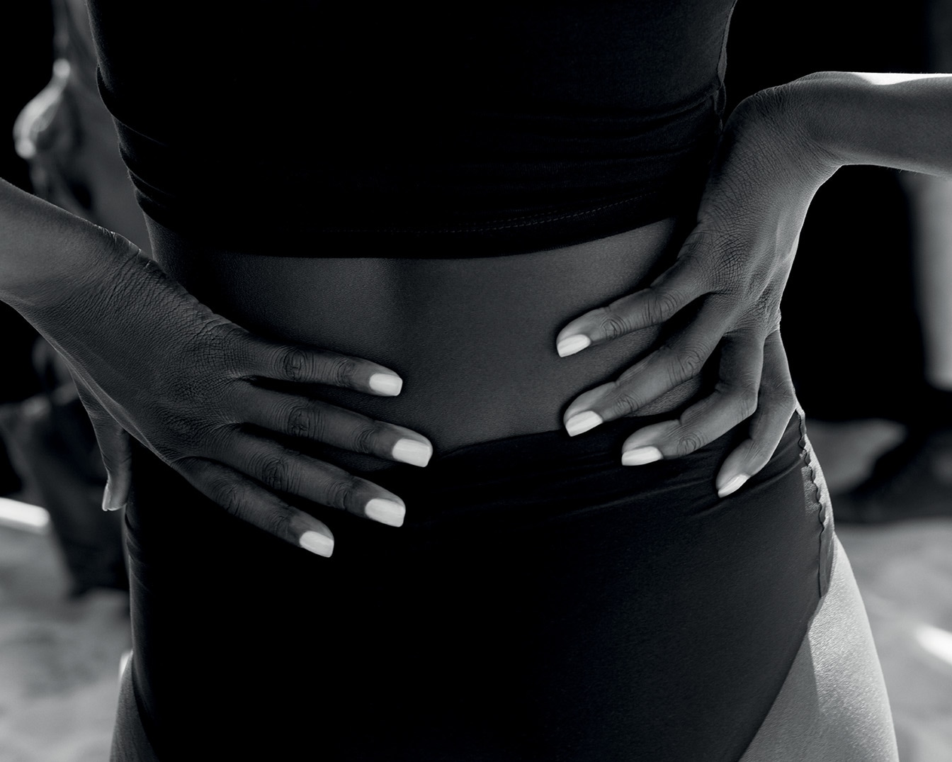 Primo piano in bianco e nero di una donna con le mani sui fianchi, indossa un top corto e slip neri, mostrando una silhouette elegante e curata.
