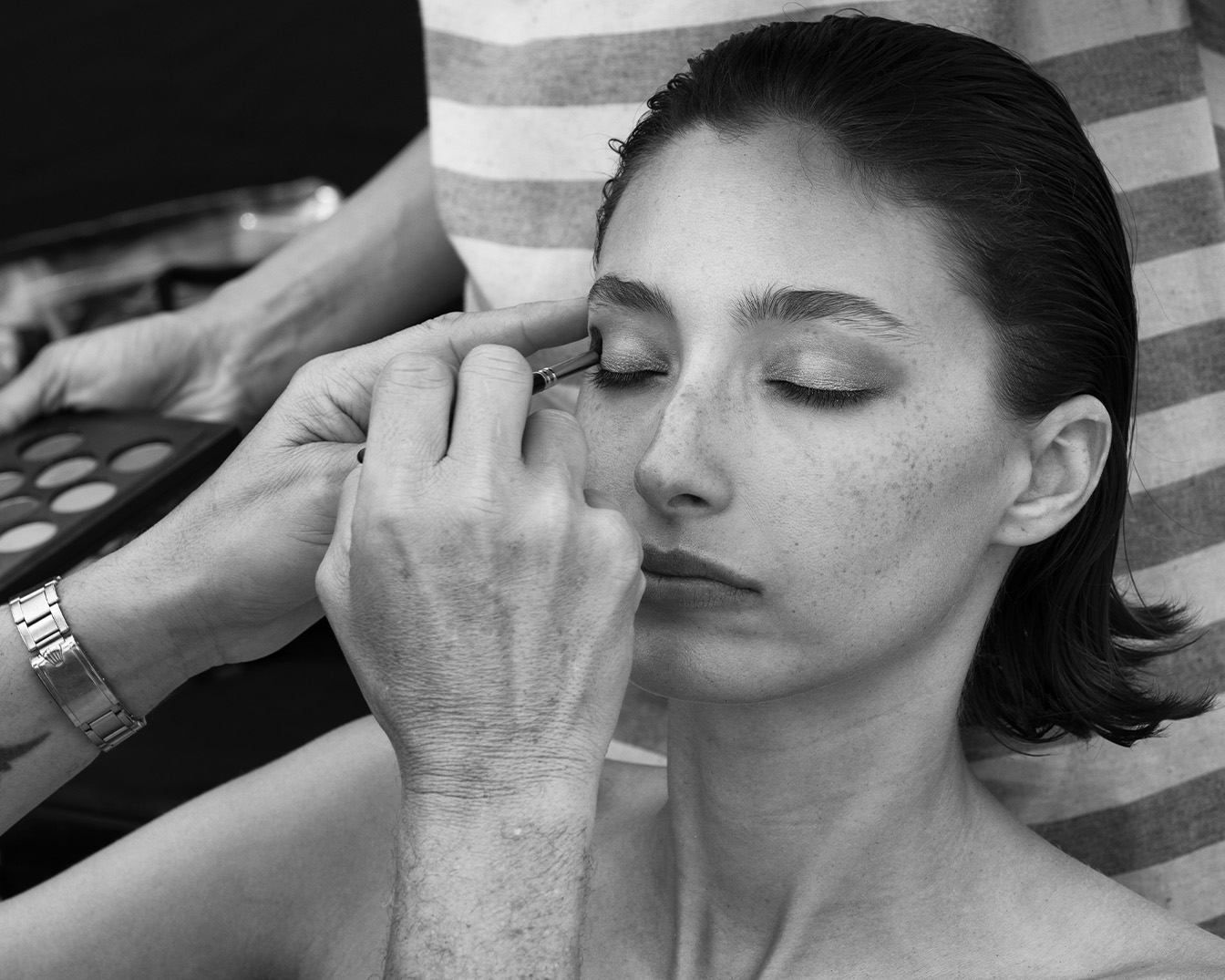 In bianco e nero, una modella con gli occhi chiusi riceve l'applicazione di ombretto da un make-up artist, che tiene in mano una palette di colori.