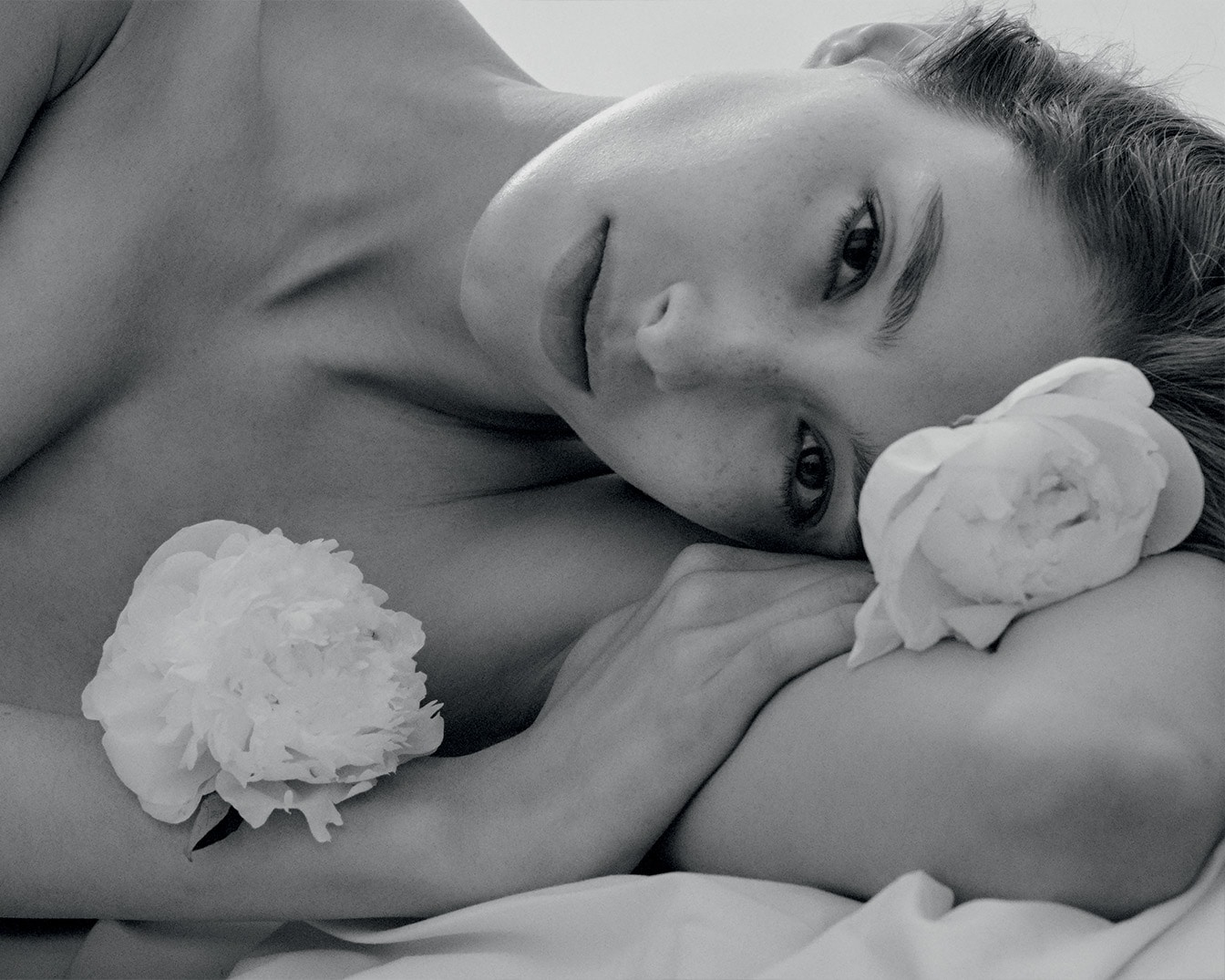 Primo piano in bianco e nero di una donna che riposa su un fianco, con fiori bianchi che le toccano la pelle, creando un'atmosfera di serena bellezza.