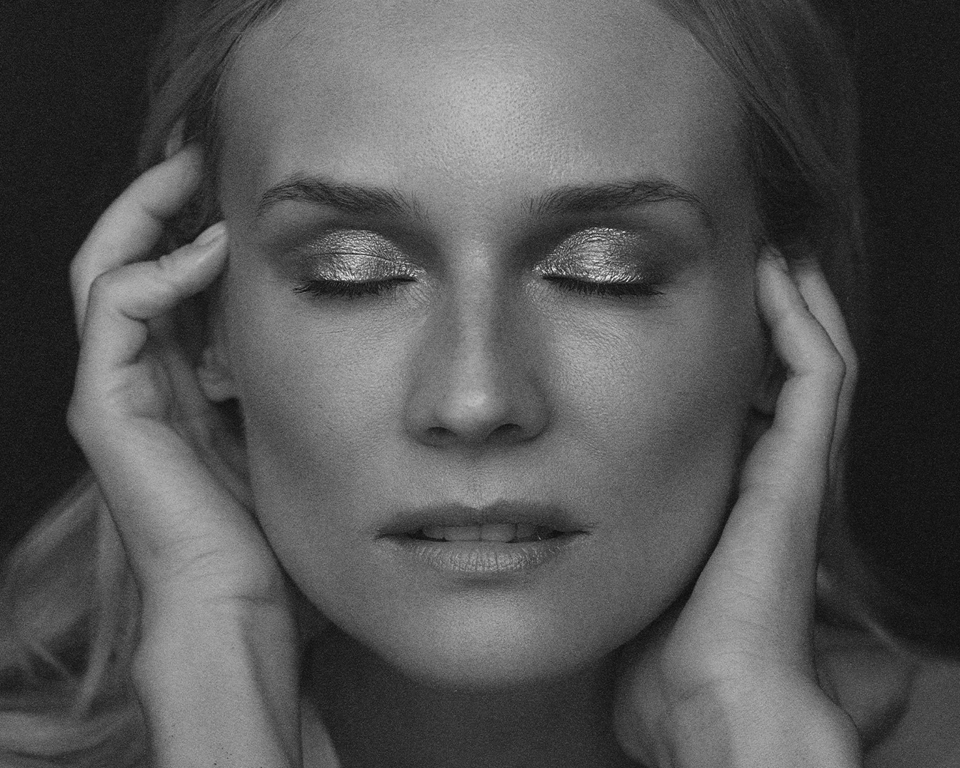 Primo piano in bianco e nero di una donna con gli occhi chiusi e ombretto argentato, le mani delicatamente appoggiate sulle tempie.