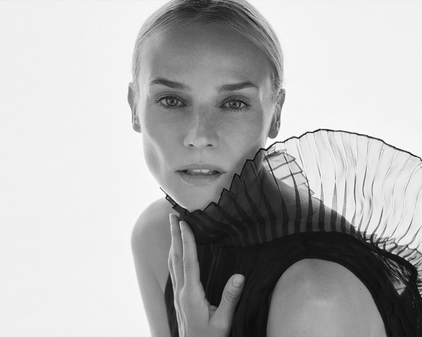 Primo piano in bianco e nero di una donna con i capelli biondi tirati indietro, indossa un abito nero con un colletto arricciato e tiene una mano delicatamente sul collo.