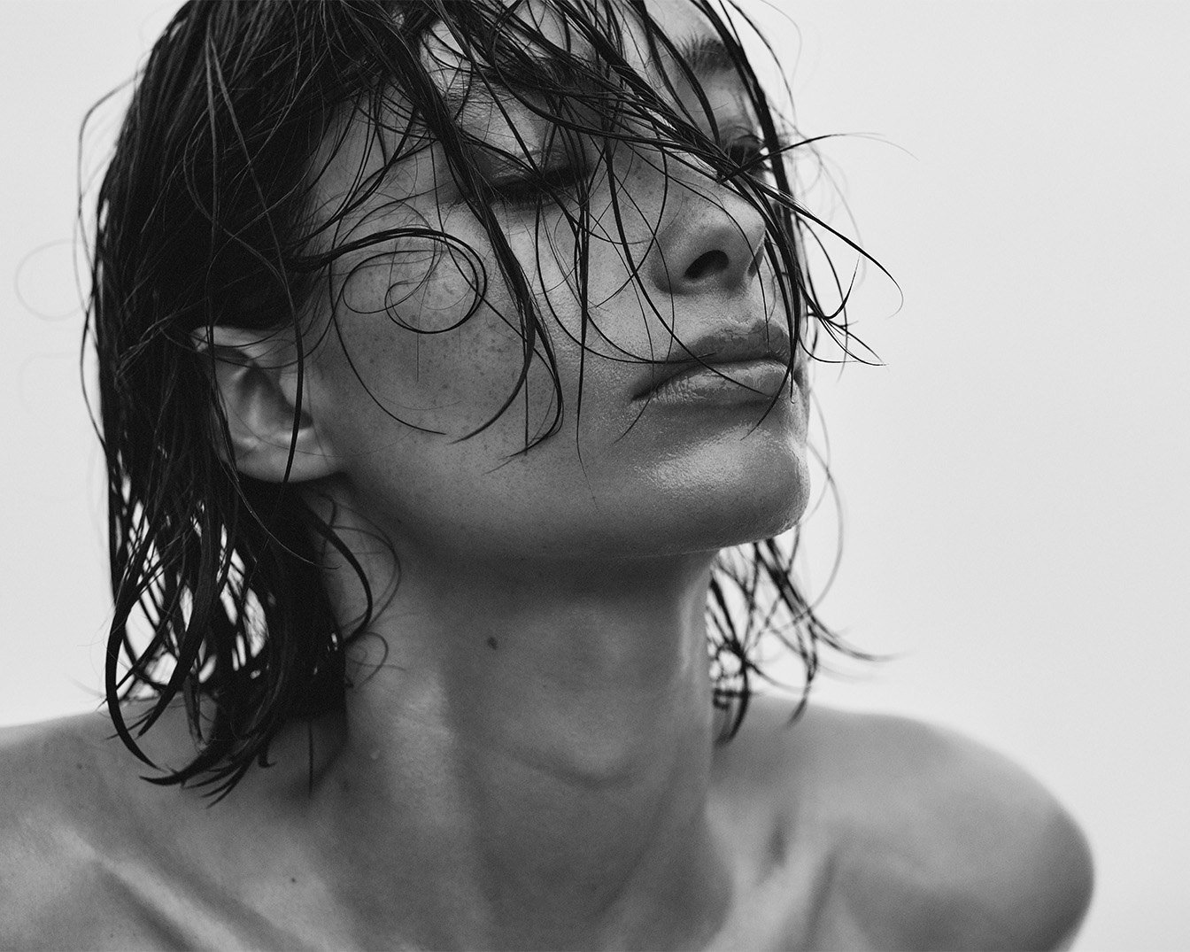 In primo piano, una donna con i capelli bagnati che le ricadono sul viso, gli occhi chiusi e la pelle luminosa, in un'immagine in bianco e nero che evoca freschezza e naturalezza.