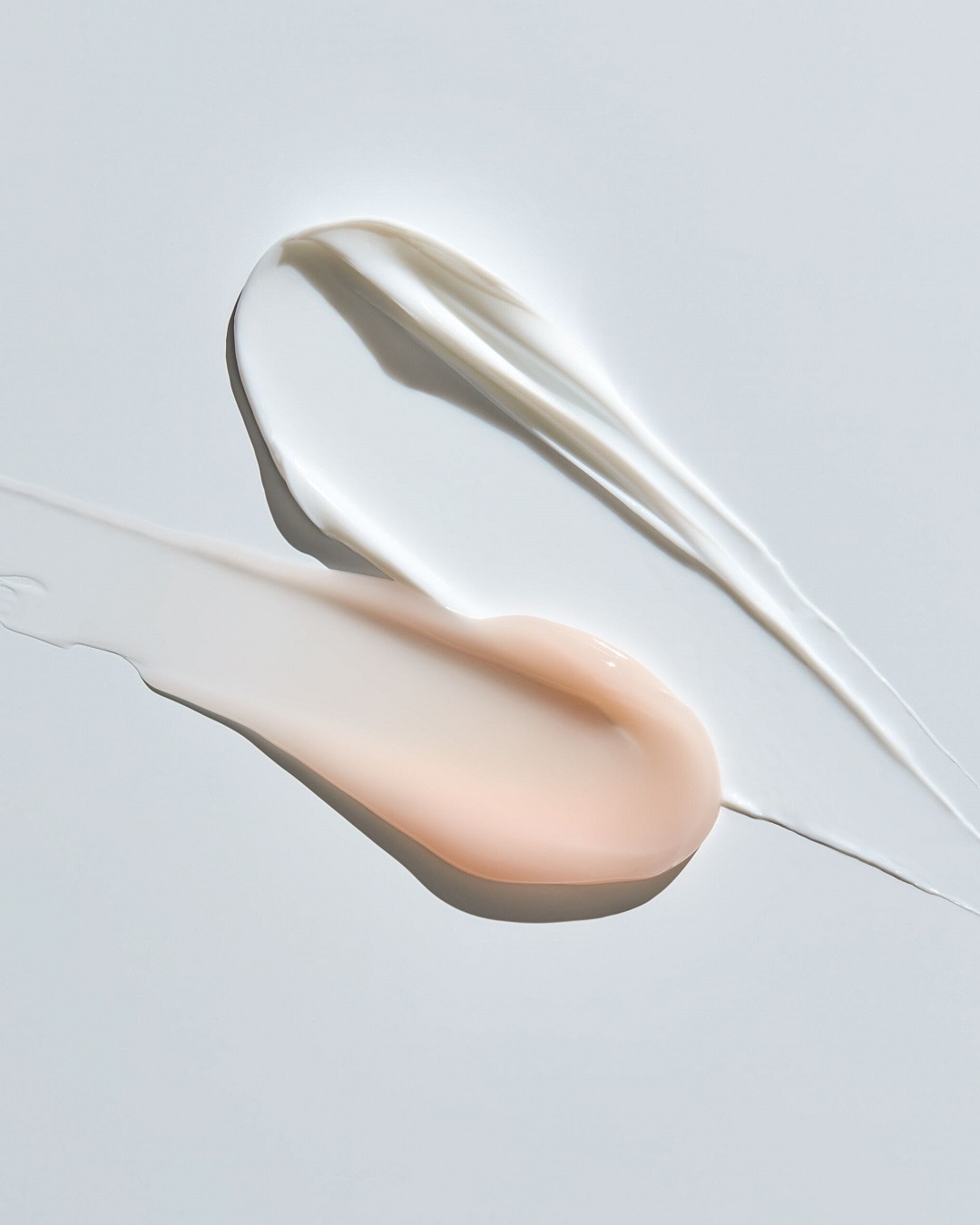 Nahaufnahme von zwei cremigen Hautpflegeprodukten, eines in Weiß und das andere in einem zarten Pfirsichton, die auf einer hellen Oberfläche aufgetragen wurden, um ihre Textur und Konsistenz zu zeigen.